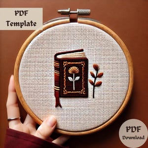 May include: Embroidery pattern for a book with a flower and a branch, in a wooden embroidery hoop. The pattern is in brown, orange, white, and cream colors. PDF Download.