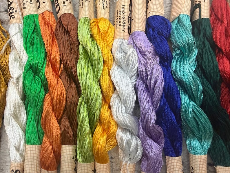 K&ouml;nnte beinhalten: Eine Sammlung farbenfroher Stickgarnkn&auml;uel. Die Garne sind in verschiedenen Farbt&ouml;nen erh&auml;ltlich, darunter Wei&szlig;, Gr&uuml;n, Orange, Braun, Gelb, Silber, Lila, Blau, T&uuml;rkis, Dunkelgr&uuml;n und Rot. Jedes Kn&auml;uel ist mit dem Wort "Embroidery Thread Fast Colour" beschriftet.