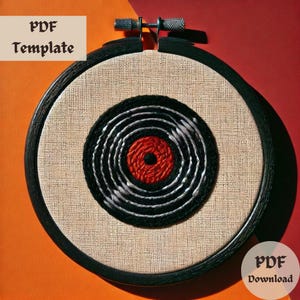 May include: A black embroidery hoop with a white and black concentric circle design and a red center. The embroidery hoop is on a red and orange background.