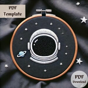 May include: Embroidered astronaut helmet design on a black fabric background with white stars and a blue planet. The design is in a round wooden embroidery hoop.