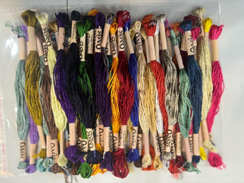 May include: A collection of 30 skeins of embroidery floss in various colors, including green, blue, red, yellow, and brown. Each skein is labeled "Oasis" and is wrapped in a white paper with a brown label.