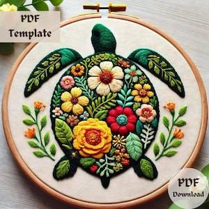 Puede incluir: Una tortuga verde con un diseño floral bordado en su caparazón. La tortuga está rodeada de hojas verdes y pequeñas flores naranjas. El bordado se realiza sobre una tela blanca y está enmarcado en un aro de madera.