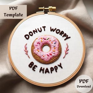 Könnte beinhalten: Stickrahmen-Kunst mit der Aufschrift "Donut Worry Be Happy" und einem rosafarbenen Donut mit Streuseln. Der Rahmen ist aus Holz und hat einen weißen Stoffhintergrund.
