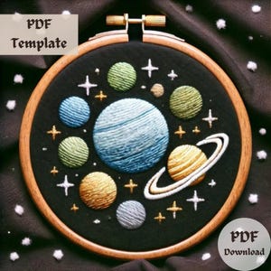 Pode incluir: Arte de bordado em aro que apresenta uma cena celestial com planetas, estrelas e um planeta anelado sobre um fundo preto. O aro é feito de madeira e tem um acabamento natural.