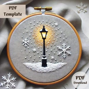 Puede incluir: Una plantilla PDF para un patrón de punto de cruz que presenta una farola con una luz brillante y copos de nieve cayendo a su alrededor. El diseño está sobre un fondo gris y está enmarcado en un aro de bordado de madera.