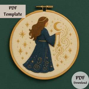 May include: Embroidered hoop art featuring a woman in a blue gown with a crescent moon and stars in gold thread. The design is on a cream-colored fabric.