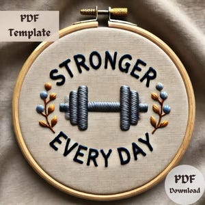Puede incluir: Aro de bordado con un fondo de tela beige y el texto "Stronger Every Day" en negro. Una mancuerna plateada está bordada en el centro del aro. Dos pequeñas ramas de flores azules y naranjas están bordadas a cada lado de la mancuerna.