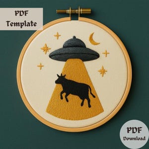 May include: Embroidered hoop art featuring a black cow being abducted by a UFO. The UFO is grey and has a yellow beam of light shining down on the cow. The background is a cream colour with yellow stars.