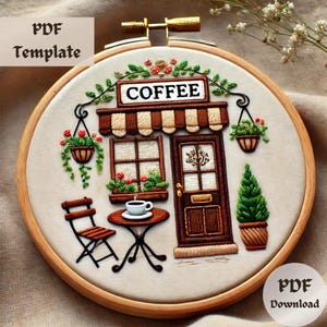 May include: Embroidered hoop art depicting a coffee shop with a brown door, a window with flowers, and a striped awning. The shop has a sign that says "COFFEE". There is a table and chair outside the shop with a cup of coffee on the table.