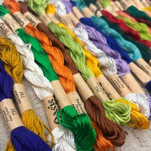 K&ouml;nnte beinhalten: Eine Sammlung farbenfroher Stickgarnspulen in verschiedenen Farbt&ouml;nen, darunter Blau, Gr&uuml;n, Orange und Gelb. Jede Spule ist mit "Embroidery Thread Fast Colour" und "TM" beschriftet. Die F&auml;den sind ordentlich angeordnet und zeigen ein Spektrum an Farbt&ouml;nen.