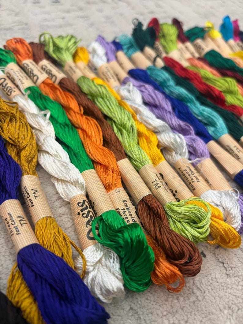 K&ouml;nnte beinhalten: Eine Sammlung farbenfroher Stickgarnspulen in verschiedenen Farbt&ouml;nen, darunter Blau, Gr&uuml;n, Orange und Gelb. Jede Spule ist mit "Embroidery Thread Fast Colour" und "TM" beschriftet. Die F&auml;den sind ordentlich angeordnet und zeigen ein Spektrum an Farbt&ouml;nen.