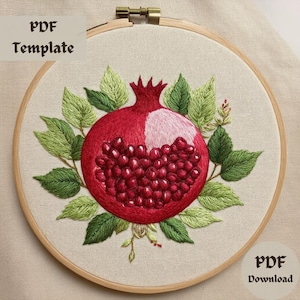 Puede incluir: Diseño bordado de una granada roja con hojas verdes sobre un fondo de tela blanca. El diseño está en un aro de bordado de madera.