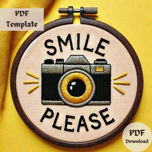 Puede incluir: Arte de bordado en aro con una cámara en blanco y negro con una lente dorada y el texto "Smile Please" en negro.