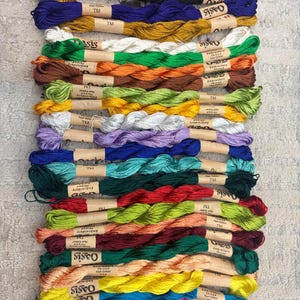 K&ouml;nnte beinhalten: Eine Sammlung farbenfroher Stickgarnkn&auml;uel in verschiedenen Farbt&ouml;nen, darunter Lila, Schwarz, Blau, Gold, Wei&szlig;, Gr&uuml;n, Braun und Gelb. Jedes Kn&auml;uel ist mit "Oasis Embroidery Thread" beschriftet.