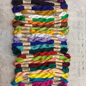 K&ouml;nnte beinhalten: Eine Sammlung farbenfroher Stickgarnkn&auml;uel, vertikal angeordnet. Die F&auml;den sind in verschiedenen Farben erh&auml;ltlich, darunter Lila, Schwarz, Blau, Gold, Gr&uuml;n, Orange, Gelb und Rosa. Jedes Kn&auml;uel hat ein Papieretikett mit der Aufschrift "Oasis Embroidery Thread TM".
