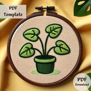 May include: Embroidered hoop art featuring a green potted plant with four leaves. The plant is in a dark green pot with a light green rim. The background is a light beige fabric.