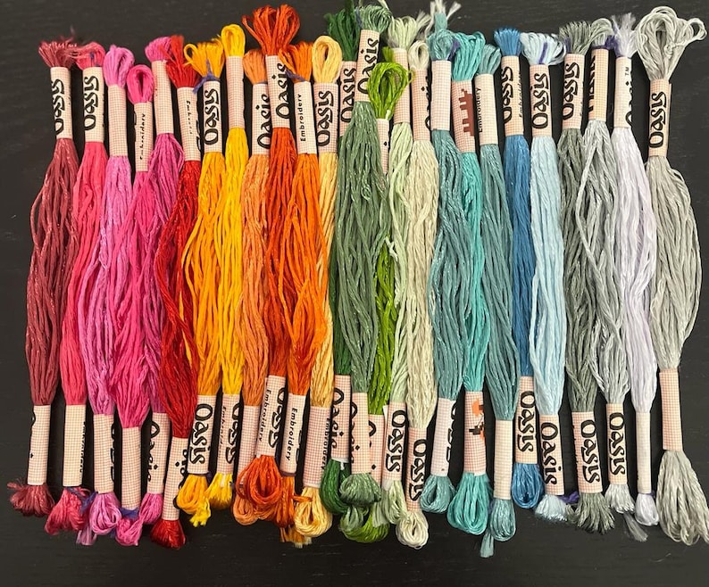 Puede incluir: Una colecci&oacute;n de 20 ovillos de hilo para bordar en varios colores, incluyendo rojo, rosa, naranja, amarillo, verde, azul y morado. Cada ovillo est&aacute; etiquetado con "Oasis" y tiene un n&uacute;mero de color diferente.