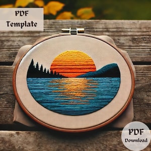 May include: Embroidered hoop art featuring a sunset over a lake with a mountain range in the background. The sunset is a gradient of orange, yellow, and red, with a blue lake reflecting the colors. The mountain range is dark blue and the trees are dark green.