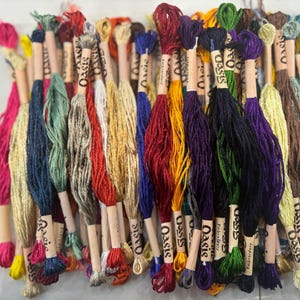 May include: A collection of colorful embroidery floss spools with the word "Oasis" printed on the labels. The floss is arranged in a variety of colors, including red, green, blue, yellow, and purple.