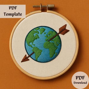 Könnte beinhalten: Stickrahmen-Kunst mit Stickerei, die einen blauen und grünen Globus mit einem braunen Pfeil zeigt, der ihn durchbohrt. Der Rahmen ist aus Naturholz und hat einen weißen Stoffhintergrund.