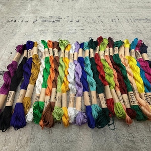 K&ouml;nnte beinhalten: Eine Sammlung bunter Stickgarne, jeweils mit einem Papieretikett mit der Aufschrift "Oasis". Das Garn ist f&auml;cherf&ouml;rmig angeordnet und zeigt eine Vielzahl von Farben, darunter Lila, Blau, Gr&uuml;n, Gelb und Orange. Ideal f&uuml;r Handarbeiten.