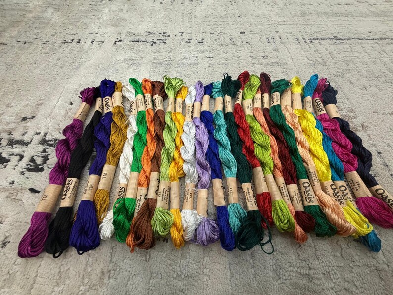 K&ouml;nnte beinhalten: Eine Sammlung bunter Stickgarne, jeweils mit einem Papieretikett mit der Aufschrift "Oasis". Das Garn ist f&auml;cherf&ouml;rmig angeordnet und zeigt eine Vielzahl von Farben, darunter Lila, Blau, Gr&uuml;n, Gelb und Orange. Ideal f&uuml;r Handarbeiten.