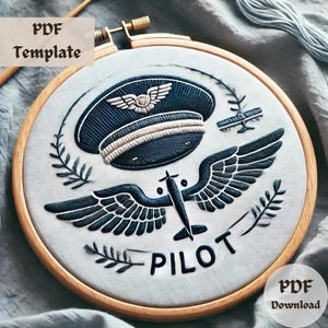 May include: Embroidered hoop art featuring a pilot's hat, wings, and airplane with the word "PILOT" below. The design is stitched in black thread on a white fabric background.