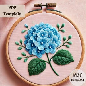 May include: A blue hydrangea flower embroidery pattern in a wooden embroidery hoop. The pattern includes green leaves and stems. PDF Download.