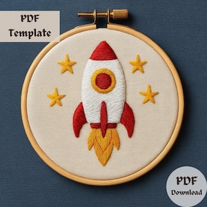 May include: Embroidered rocket ship design in a wooden hoop. The rocket is white with red accents and yellow flames, surrounded by yellow stars. The text "PDF Template" and "PDF Download" are visible.