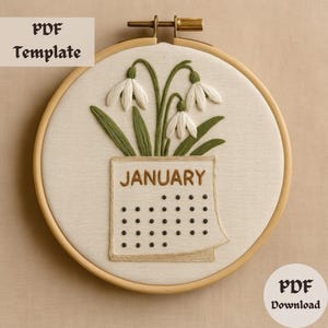 Op de afbeelding: Borduurwerk in een borduurring met een wit sneeuwklokje-boeket met groene bladeren en een kalender met het woord "JANUARY" en zwarte stippen voor de dagen van de maand.