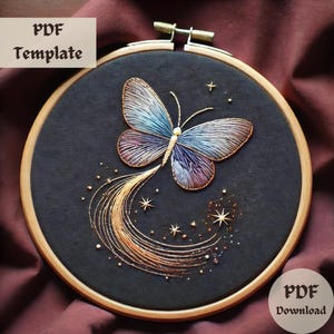 May include: A black embroidery hoop featuring a colourful butterfly design with gold glitter accents. The butterfly has a long tail of gold glitter trailing behind it. The hoop is on a burgundy fabric background.