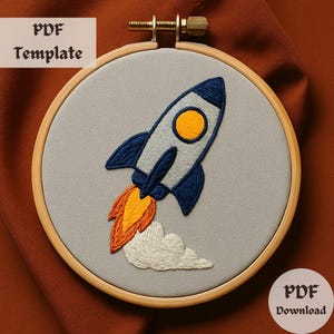 May include: A PDF template for embroidery featuring a rocket ship with orange and yellow flames and a white smoke trail. The rocket is blue and gray with a yellow circle in the middle. The template is in a wooden embroidery hoop.