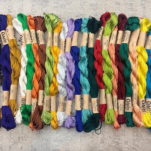 K&ouml;nnte beinhalten: Eine Sammlung farbenfroher Stickgarne in verschiedenen Farbt&ouml;nen, darunter Lila, Schwarz, Wei&szlig;, Gelb, Orange, Gr&uuml;n und Blau. Jedes Kn&auml;uel ist mit einem Papieretikett umwickelt. Die Garne liegen auf einer hellgrauen Oberfl&auml;che.