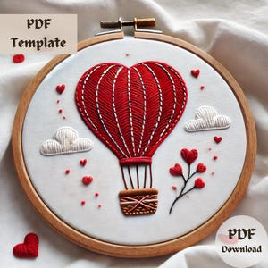 May include: Embroidered hoop art featuring a red heart-shaped hot air balloon with white clouds and red hearts scattered around it. The balloon has a basket with a brown and white pattern. The hoop is made of wood and has a white background.