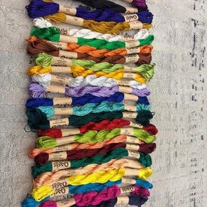 K&ouml;nnte beinhalten: Eine Sammlung farbenfroher Stickgarne, jeweils mit einem Papieretikett umwickelt. Die Garnfarben umfassen Schattierungen von Lila, Schwarz, Blau, Gold, Gr&uuml;n, Orange, Braun, Gelb und Rosa. Der Markenname "Oasis" ist auf den Etiketten sichtbar.