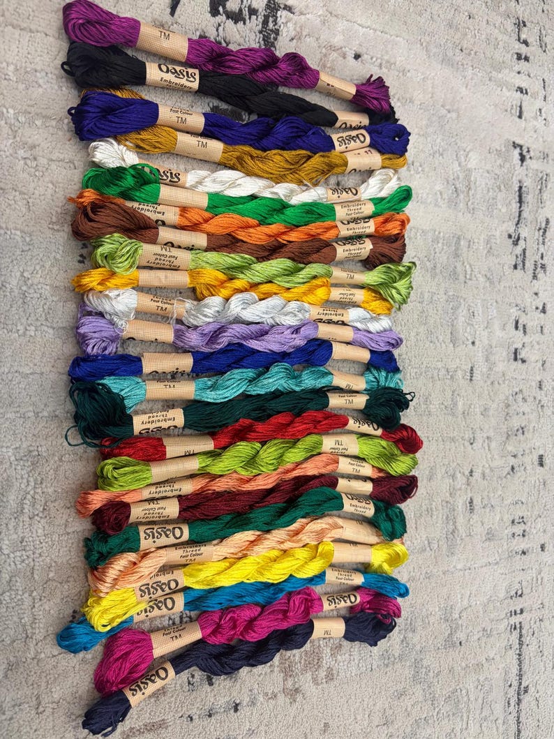 K&ouml;nnte beinhalten: Eine Sammlung farbenfroher Stickgarne, jeweils mit einem Papieretikett umwickelt. Die Garnfarben umfassen Schattierungen von Lila, Schwarz, Blau, Gold, Gr&uuml;n, Orange, Braun, Gelb und Rosa. Der Markenname "Oasis" ist auf den Etiketten sichtbar.