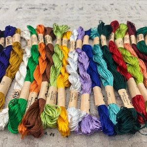 K&ouml;nnte beinhalten: Eine Sammlung farbenfroher Stickgarne in verschiedenen Farbt&ouml;nen, darunter Lila, Schwarz, Gelb, Gr&uuml;n, Orange und Blau. Jedes Kn&auml;uel ist mit einem Papieretikett umwickelt. Die F&auml;den sind eng aneinander angeordnet und zeigen ein Spektrum an Farbt&ouml;nen.