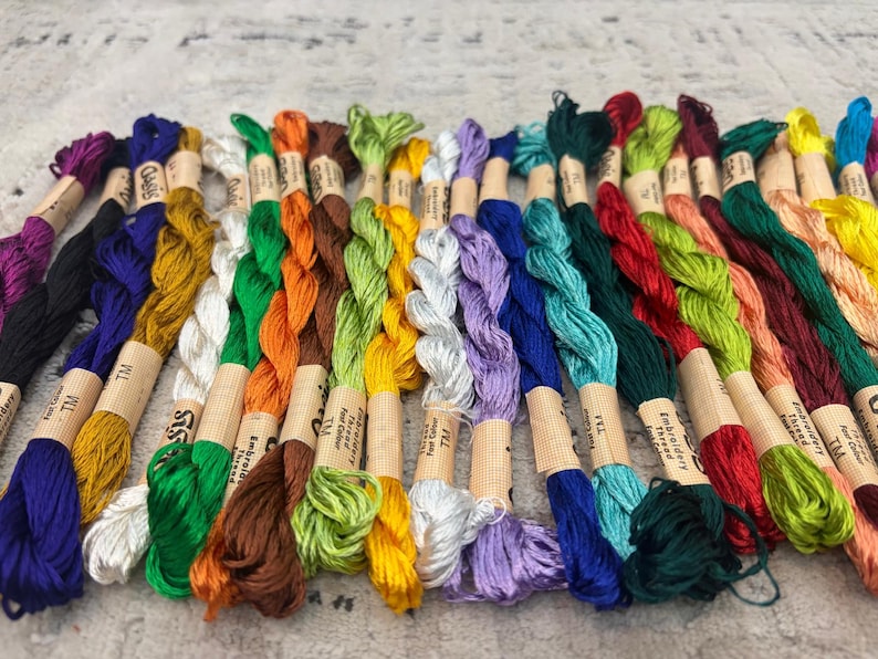 K&ouml;nnte beinhalten: Eine Sammlung farbenfroher Stickgarne in verschiedenen Farbt&ouml;nen, darunter Lila, Schwarz, Gelb, Gr&uuml;n, Orange und Blau. Jedes Kn&auml;uel ist mit einem Papieretikett umwickelt. Die F&auml;den sind eng aneinander angeordnet und zeigen ein Spektrum an Farbt&ouml;nen.