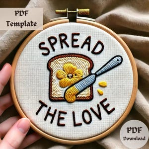 May include: Embroidered hoop art with the words "SPREAD THE LOVE". The design features a slice of toast with a flower and a butter knife. The embroidery is in shades of brown, yellow, and white, set against a cream-colored fabric.