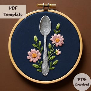 May include: A silver spoon with a pink flower embroidery design on a blue background. The embroidery is in a hoop with a wooden frame.