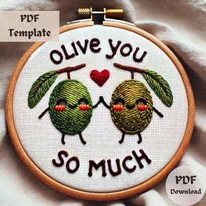 Puede incluir: Arte de bordado en un aro con dos aceitunas verdes que se toman de la mano y un corazón rojo entre ellas. El texto "Olive you so much" está bordado debajo de las aceitunas.