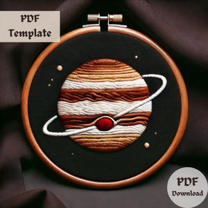 Puede incluir: Arte de bordado en aro que presenta una representación detallada de Júpiter, el planeta más grande de nuestro sistema solar. El planeta está representado en tonos marrones y blancos, con una mancha roja que representa la Gran Mancha Roja. El aro está hecho de madera y tiene un fondo de tela negro.