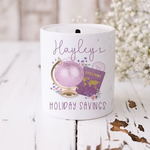May include: White ceramic money box with a pink globe, passport, and floral design. The text "Hayley's Holiday Savings" is printed on the front.