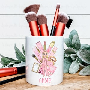 May include: A white ceramic makeup brush holder featuring a pink and gold illustration of makeup items and a girl in pyjamas. The holder contains makeup brushes with red handles and brown bristles. The name "Abbie" is printed on the front.