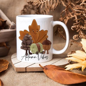 May include: White ceramic mug with an autumn-themed illustration. The design features a smiling leaf, pinecone, chestnut, and acorn, with the words "Autumn Vibes". The mug is on a wooden coaster. The mug is perfect for a warm beverage.