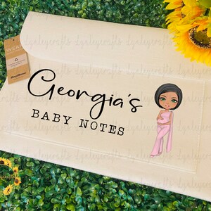 May include: A cream-coloured baby notes organiser with the text "Georgia's BABY NOTES" in black script. A cartoon illustration of a pregnant woman in pink is on the right. The organiser has a flap closure and a small pocket.