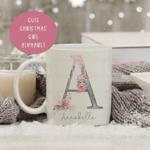 May include: White ceramic mug featuring a pink and grey letter A, a pink Christmas elf illustration, and the name Annabelle. The mug is on a white surface with a grey knitted blanket.