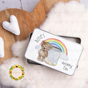 May include: A white metal tin with a rainbow, clouds, and a teddy bear holding a jar filled with stars. The tin says "Alfie's Worry Tin".