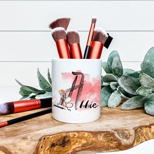 May include: White ceramic makeup brush holder with a pink watercolour design and the name "Abbie" written in black. The holder is filled with makeup brushes.