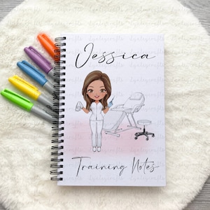 May include: A white spiral bound notebook with a pink and white gradient background. The notebook features a cartoon illustration of a woman in a white uniform holding a syringe and a medical chair. The text "Jessica" and "Training Notes" are printed on the cover.